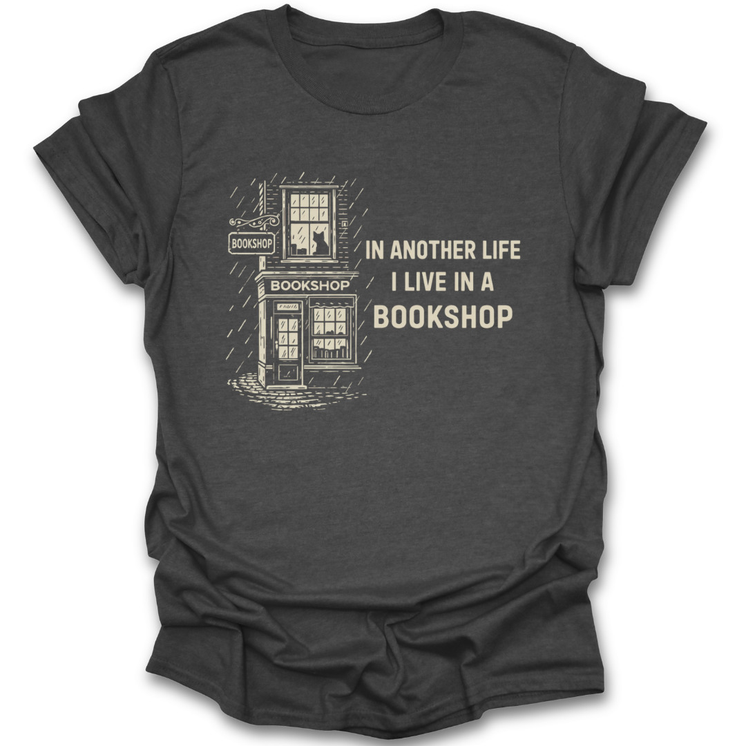 Dark gray t-shirt with a graphic of a bookshop and text 'In another life I live in a bookshop' on a white background. funny book reading literary t shirt