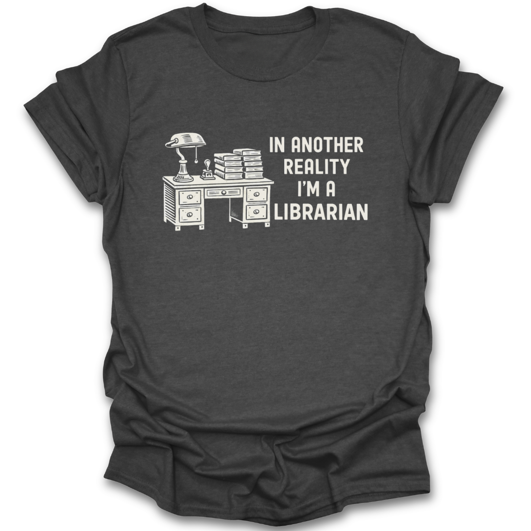 Black t-shirt with a graphic of a desk and books, and text 'In another reality I'm a librarian' on a white background. funny book reading literary t shirt