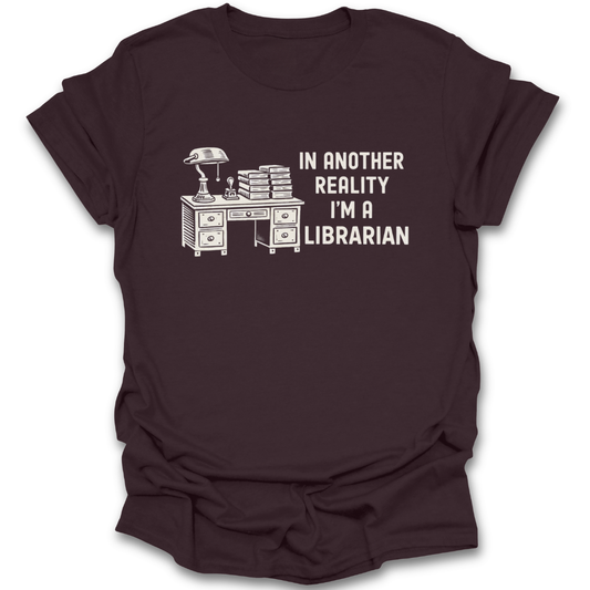Brown t-shirt with a graphic of a desk and books, and text 'In another reality I'm a librarian' on a white background. funny book reading literary t shirt