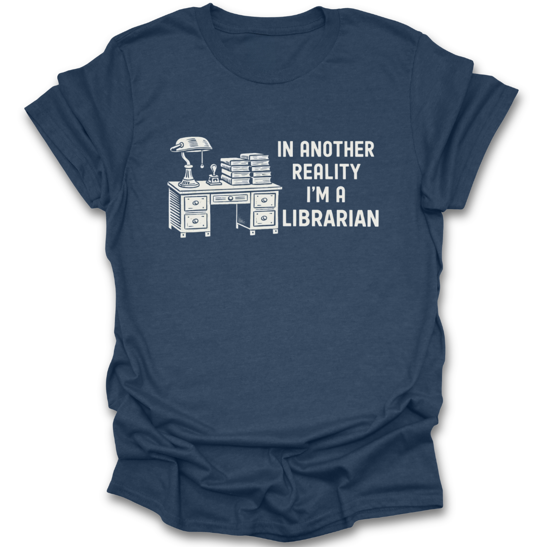 Blue t-shirt with a graphic of a desk and books, and text 'In another reality I'm a librarian' on a white background. funny book reading literary t shirt