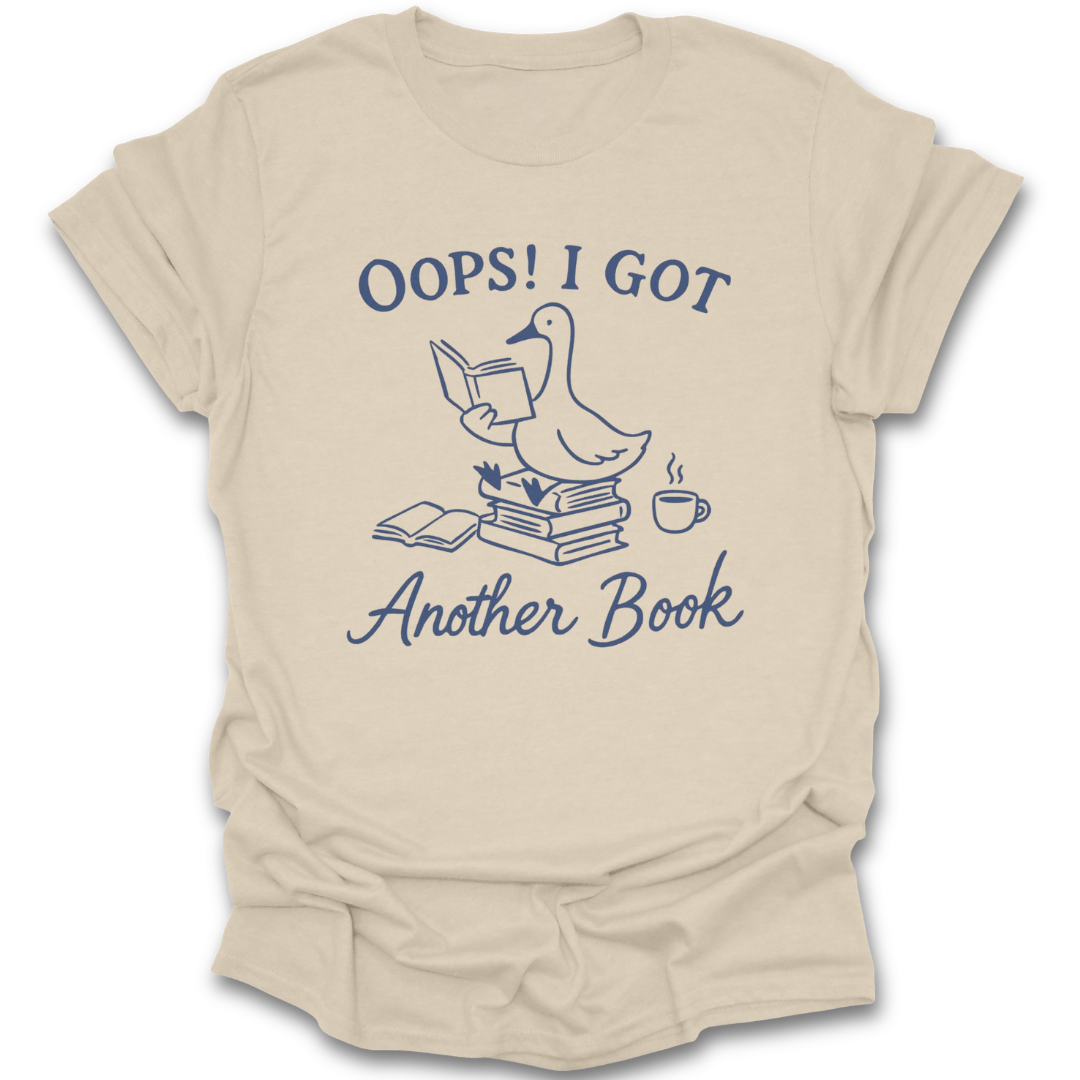 Beige t-shirt with a graphic of a duck reading a book and text 'Oops! I got another book' on a white background. oops i got another book shirt, funny book lover shirt, bookish t-shirt, reader gift, bookworm apparel, bookstore shirt, bibliophile tee, reading humor shirt, book club gift, coffee and books shirt