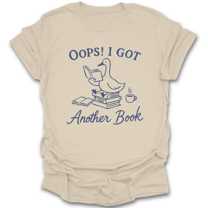 Beige t-shirt with a graphic of a duck reading a book and text 'Oops! I got another book' on a white background. oops i got another book shirt, funny book lover shirt, bookish t-shirt, reader gift, bookworm apparel, bookstore shirt, bibliophile tee, reading humor shirt, book club gift, coffee and books shirt