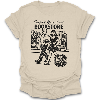 Beige t-shirt with a graphic of a man and woman walking past a bookstore and text 'Support Your Local Bookstore'. funny book reading literary t shirt