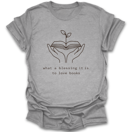 Gray t-shirt with a graphic of hands holding an open book with a plant growing out of it and text 'what a blessing it is to love books' on a white background. book reading literary t shirt