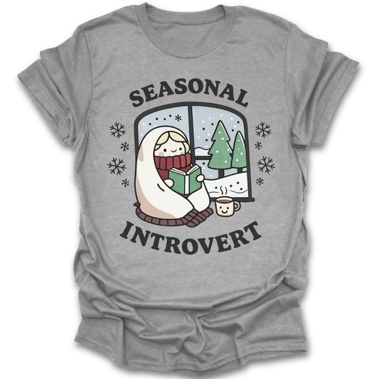Gray t-shirt with 'Seasonal Introvert' graphic design featuring a person reading a book by a window with trees. funny book reading literary t shirt